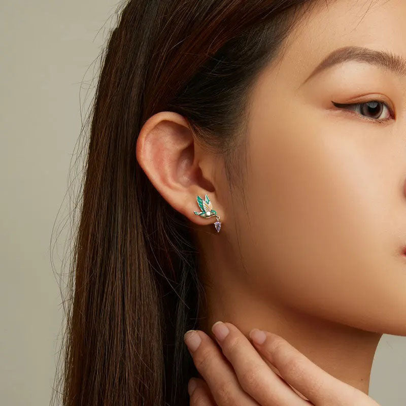 Close-up of a sterling silver hummingbird stud earring, featuring vibrant teal and peach enamel with a dangling purple teardrop crystal, elegantly worn on a model's ear.