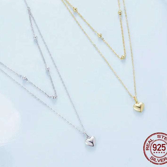 Flat lay of layered heart pendant necklaces in 925 sterling silver with gold-tone and silver finishes, beaded station and link chains on a light background — OmniVivo