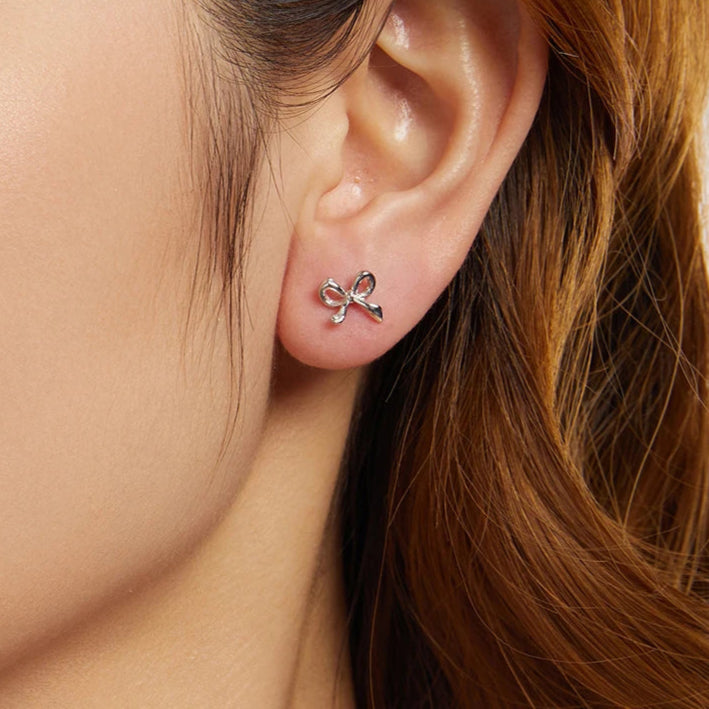 On-ear close-up of mini bow stud earring in 925 sterling silver with white-gold plating, minimalist ribbon design — OmniVivo