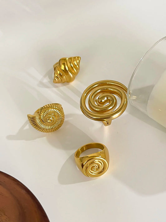 A collection of luxurious gold-toned rings and a charm from the Azure series, featuring intricate conch seashell and spiral designs, elegantly displayed on a minimalist surface to highlight their unique, ocean-inspired artistry.