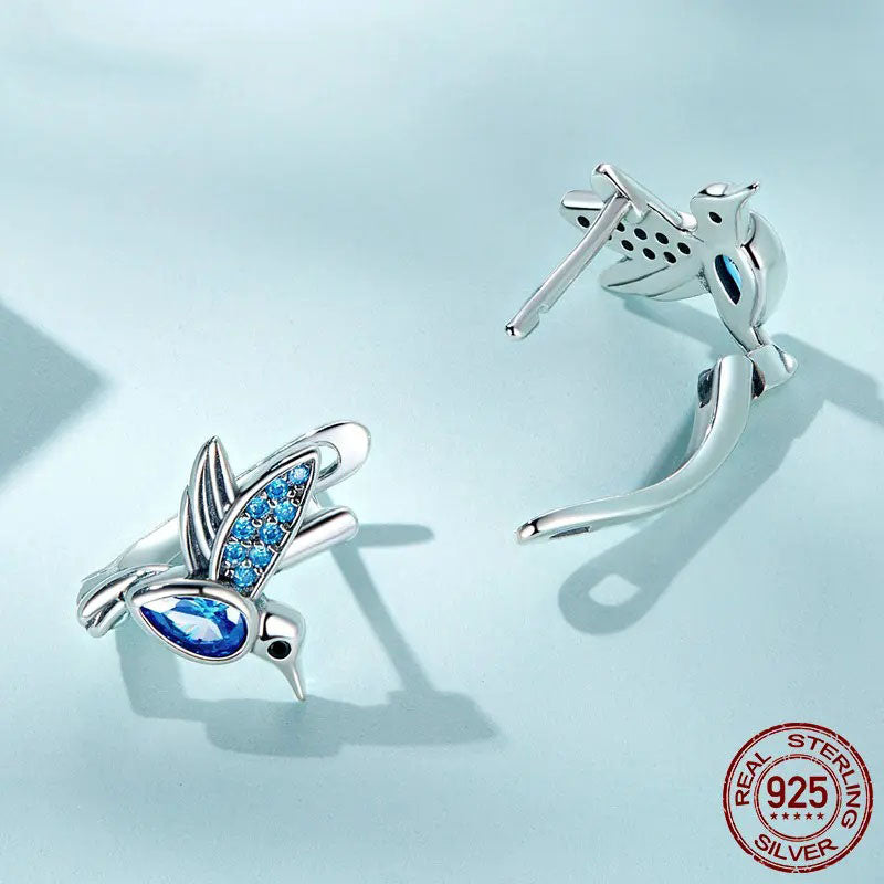 A pair of sparkling sterling silver hummingbird-shaped hoop earrings, intricately detailed with blue cubic zirconia stones on the wings and a larger blue teardrop crystal as the body, showcasing their unique and elegant design.