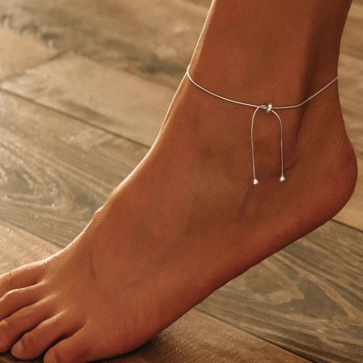 Adjustable 925 sterling silver snake chain anklet worn on the ankle, lifestyle shot on a wooden floor.