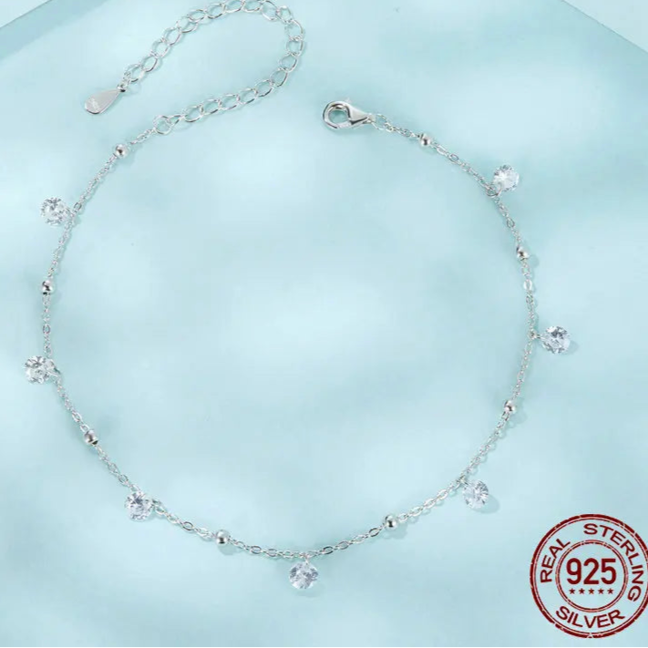Top view of a 925 sterling silver cubic zirconia anklet with adjustable extender and dangle charms, arranged in a circle — light blue background with 925 stamp.