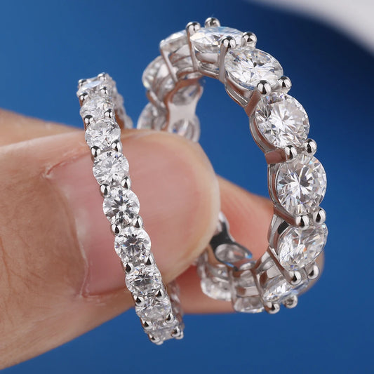 Close-up of 3 mm and 5 mm moissanite row rings in solid 925 sterling silver, prong-set eternity style, held between fingers on blue background.