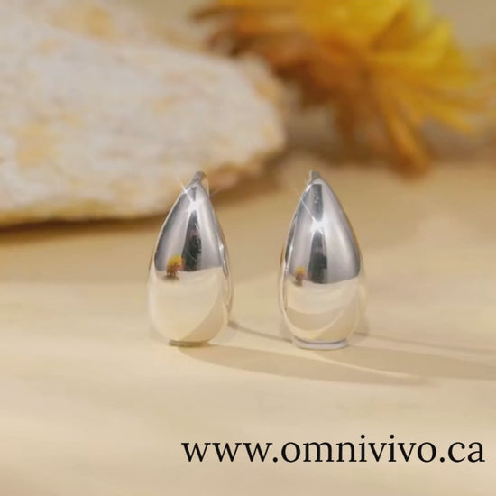 Glossy teardrop earrings in 925 sterling silver, 15×8 mm waterdrop design, shown on a warm floral backdrop — OmniVivo