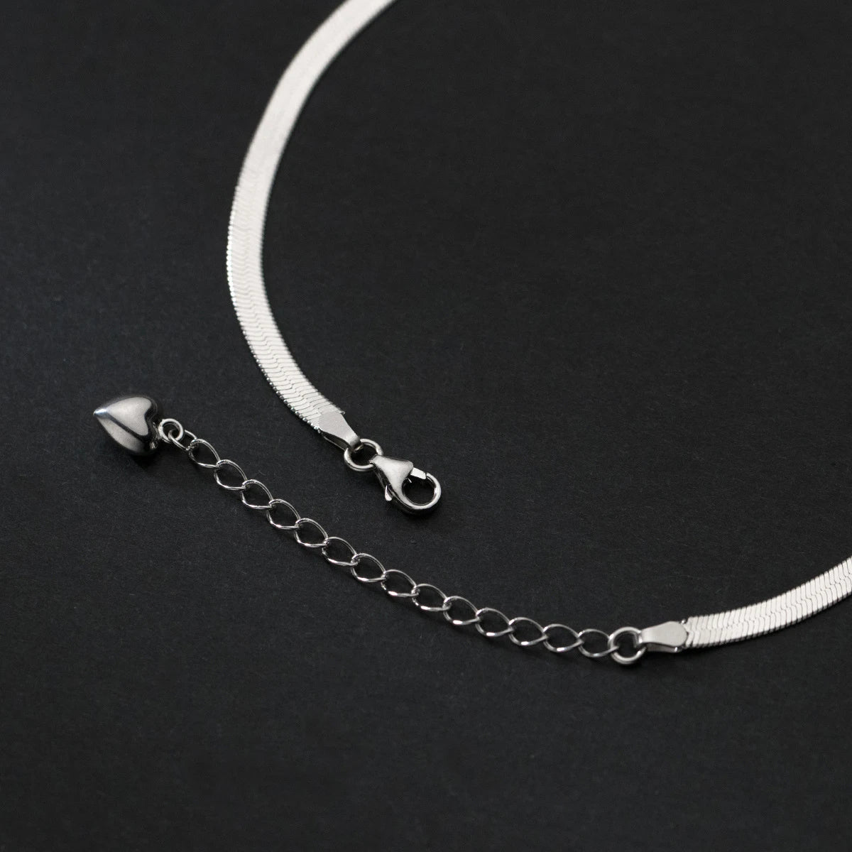 Silver-tone snake chain anklet with lobster clasp and extender, heart end charm, on a black background.